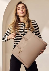 Beige leather tote bag with textured surface, flat shape, and golden hardware detail. Model holds it on a neutral background.