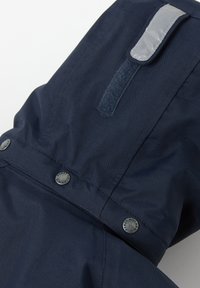 Meriveden kestävä, vettä hylkivä takki, jossa on rakenteellinen kangas, harmaat heijastavat yksityiskohdat, napit ja kohotettu kaulus.