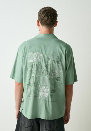 Mann trägt ein hellgrünes Kurzarmhemd mit detaillierten botanischen Illustrationen und Text auf dem Rücken.