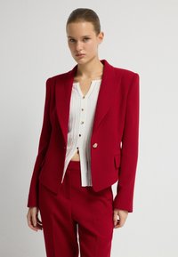 Červený přizpůsobený blejzr z jednolité tkaniny; ostré klopy, jednobotonové zapínání a dvě boční kapsy. Noste ho přes bílou skládací košili.