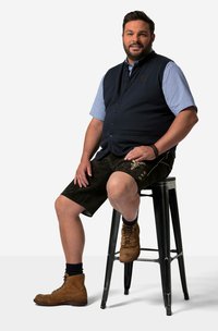 Man sitter på en svart pall, iförd en mörkblå väst över en ljusblå skjorta, bruna skinnshorts och bruna snörskor med mörka strumpor.