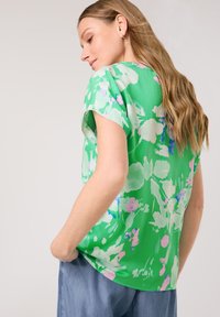 Grüne Blumenshirt mit kurzen Ärmeln, mit pinken und blauen Akzenten, weichem Material und entspanntem Schnitt, kombiniert mit hellblauen Jeans.