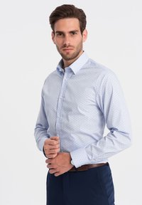 Camicia azzurro chiaro con piccoli pois scuri, design aderente, colletto alla francese e maniche lunghe. Abbinata a una cintura scura.
