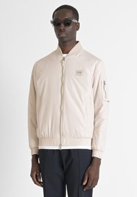 Chaqueta bomber beige claro con cierre frontal, puños acanalados, bolsillos laterales y un parche con logo en el pecho. Tejido suave y brillante con una silueta ajustada.