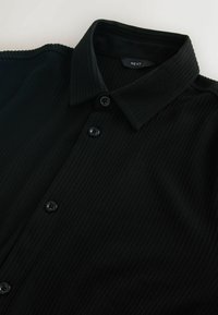 Melnas pogājamas krekls, kas izgatavots no ribota auduma. Tam ir klasiskā apkakle un piecas melnas pogas priekšpusē. Vienkāršs, elegants dizains.