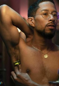 Bare-chested man with dark hair, wearing clear glasses and a gold necklace, shaving armpit with a green and brown razor.
