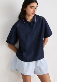 Polo shirt blu navy con colletto a righe bianche e rosse, caratterizzato da pannelli laterali in rete; abbinato a pantaloni corti a righe blu chiaro.