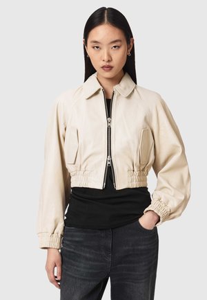 Młoda kobieta ubrana w krótką kremową kurtkę na suwak z elastycznymi mankietami i krawędzią, czarną koszulę i ciemne dżinsy, stojąca na jednolitym tle.