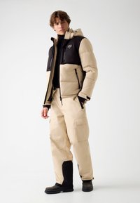 Beige Daunenjacke mit schwarzen Schultern, Kapuze und Reißverschlusstaschen, kombiniert mit beige Cargo-Hosen mit großen Seitentaschen und schwarzen Akzenten.