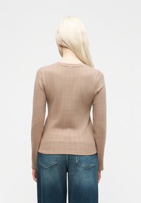 Ξανθό άτομο που φοράει εφαρμοστή, μακρυμάνικη μπεζ μπλούζα με διακριτικά κάθετα σχέδια και φαρδύ μπλε τζιν, γυρισμένο πλάτη σε λευκό φόντο.