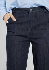 Ciemnoniebieskie dżinsy o slim fit, z pięcioma kieszeniami oraz metalowymi guzikami i nity. Gładka tekstura z subtelnym stretch.