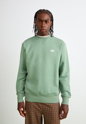 Mint green sweatshirt with a crew neck, ribbed cuffs, and a small logo on the chest. Soft fabric with a relaxed fit, paired with plaid trousers.