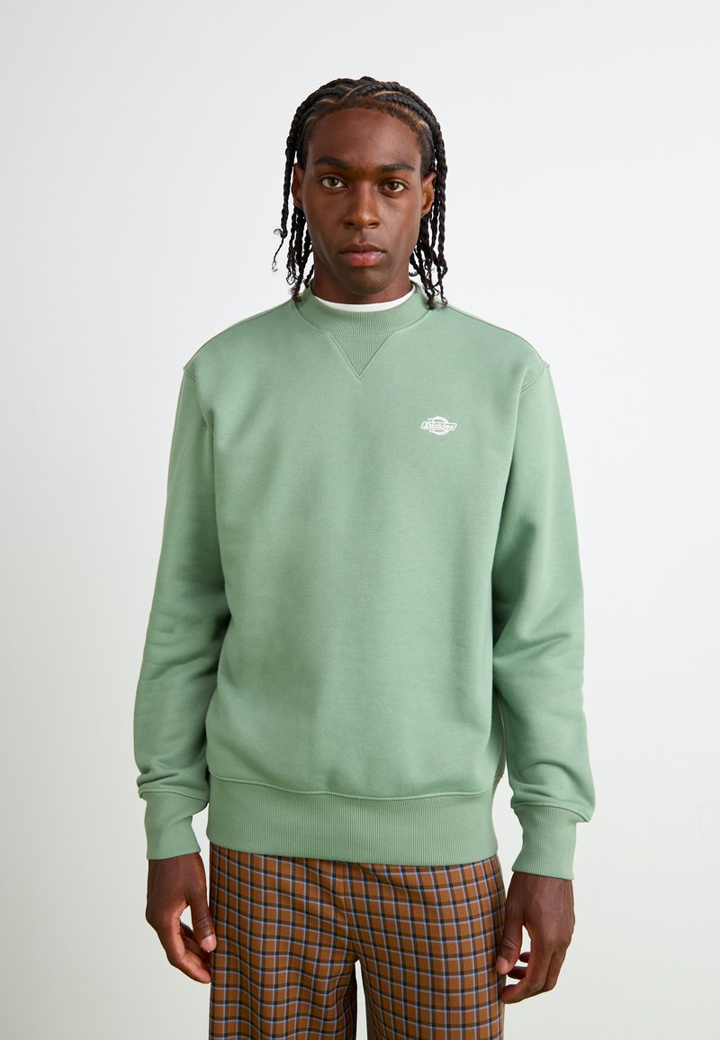 Mintgroene sweatshirt met ronde hals, geribbelde mouwen en een klein logo op de borst. Zacht materiaal met een relaxte pasvorm, gecombineerd met een plissébroek.