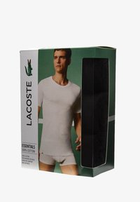 Il packaging degli slip neri Lacoste da uomo mostra una figura che indossa una t-shirt bianca in cotone. La scatola presenta uno sfondo verde e il logo.