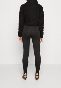 Schwarzer, kabelgestrickter, verkürzter Pullover mit hohem Kragen, kombiniert mit körperbetonten schwarzen Ripp-Leggings mit Knöchelausschnitten und Absatzsandalen.