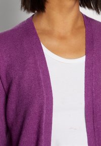 Fialový cardigan z jemného materiálu s pohodlným střihem a otevřeným předním dílem. Nosený přes bílý tričko s kulatým výstřihem.