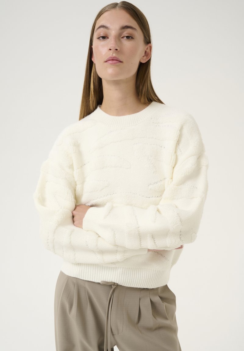 Cremfarbener Strickpullover mit strukturierten Mustern, Rundhalsausschnitt und gerippten Bündchen, kombiniert mit hellbeigen Hosen. Schlichtes, gemütliches Design.