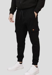 Pantaloni jogger neri con tasche cargo laterali, polsini elasticizzati e vita con cordoncino, abbinati a sneakers bianche. Presenta un patch con logo colorato.
