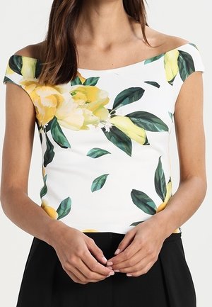 Biała bluzka z odkrytymi ramionami, ozdobiona żółtymi kwiatowymi wzorami i zielonymi liśćmi, o dopasowanym kroju i gładkiej fakturze, w zestawieniu z czarnymi spodnimi.