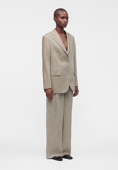 Femme portant un blazer beige oversize et un pantalon large avec des chaussures noires pointues, debout devant un fond blanc uni.