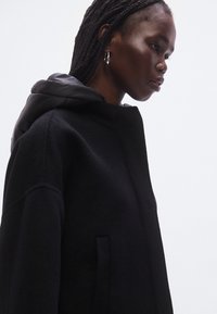 Manteau noir avec un extérieur en laine texturée et une capuche matelassée contrastante. Doté d'un col montant et de poches latérales discrètes.