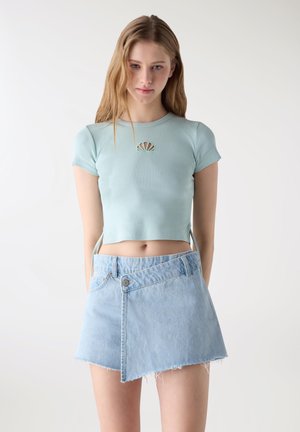 Giovane donna che indossa un top corto azzurro chiaro con ritaglio a conchiglia e una minigonna di jeans chiaro asimmetrica, in piedi contro uno sfondo bianco semplice.