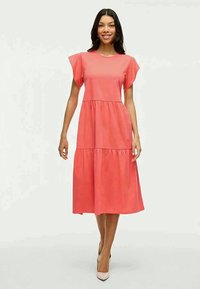 Vestido cor-de-coral, até aos joelhos, com mangas curtas em estilo de asas, design em camadas e textura suave, combinado com sapatos de salto neutros.