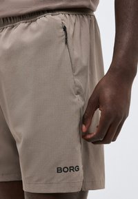 Μπεζ σορτς με μικρό grid σχέδιο, τσέπη με φερμουάρ κοντά στη μέση και μαύρο λογότυπο "BORG" κοντά στο στρίφωμα στο αριστερό πόδι, φορεμένα από άτομο με σκούρο δέρμα.