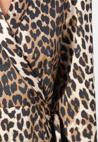 Leopardprint omslagstop i silkeagtigt stof. Har en tanfarvet base med sorte og brune pletter. Bundet om taljen, har den en glat tekstur og blød fald.