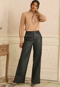 Beige džemperis ar teksturētām piedurknēm un loku ap kaklu, apvienots ar augstā jostasvietā melniem mākslīgās ādas biksēm ar pogu detaļām.