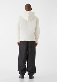 Krémszínű kapucnis pulóver nagy kapucnival, laza szabású, sötét farmernadrággal kombinálva, hátsó zsebekkel. Sima textúra, minimalistába tervezés.