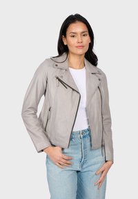 Giacca da motociclista in pelle grigia con un design aderente, spalline sulle spalle e numerosi zip. Indossata sopra una maglietta bianca e jeans in denim.