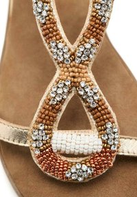 Sandalo con perline caratterizzato da una fascetta dorata, un intricato motivo di perline in tonalità marrone, bianco e arancione, e decorazioni in cristallo su un sottopiede beige.