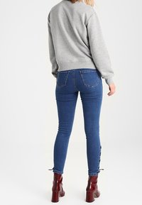 Sudadera gris con escote redondo, combinada con vaqueros ajustados azules que presentan detalles de botones en el tobillo. La modelo lleva botas rojas de tobillo.