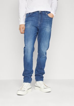 Person trägt blaue Jeans, weißes Langarmhemd und weiße Sneaker und steht mit einer Hand in der Tasche vor einem schlichten Hintergrund.