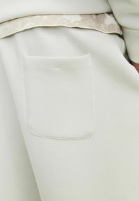 Pantaloni jogger gri deschis cu un buzunar pe spate, material moale și o textură netedă, având un brâu în contrast cu un model subtil.