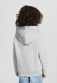 Felpa con cappuccio grigia dal morbido texture, polsini e orlo a costine. Vista posteriore che mostra una vestibilità rilassata e un design semplice. Abbinata a jeans azzurri chiari.