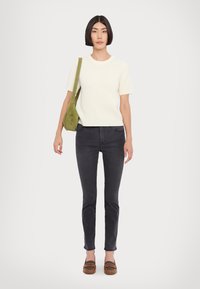 Cremefarbener, gerippter Strickpullover mit kurzen Ärmeln, kombiniert mit dunkelgrauen, schmalen Jeans und braunen Schuhen. Eine grüne Schultertasche setzt einen kontrastierenden Akzent.