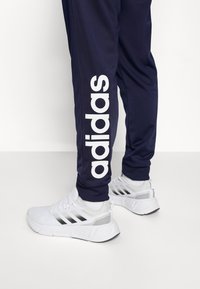 Pantalones deportivos marinos con el logo blanco de "adidas" a lo largo del costado, combinados con zapatillas blancas con detalles en gris y tres rayas negras.