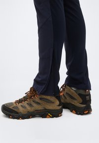 Άτομο που φορά καφέ και μαύρα παπούτσια πεζοπορίας με πορτοκαλί λεπτομέρειες και σκούρο μπλε παντελόνι με φερμουάρ, στέκεται σε γκρι επιφάνεια.