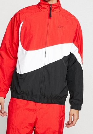 Mężczyzna w czerwonej, białej i czarnej kurtce Nike na zamek błyskawiczny oraz pasujących czerwonych spodniach, stojący na tle jednolitego jasnego tła.