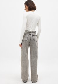 Pitkähihaisessa valkoisessa paidassa on korkeavyötäröiset, leveälahkeiset harmaat farkut, joissa on kulutettu pinta ja perinteinen viisi taskua -muotoilu.