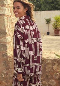 Bordeaux geklede shirt met witte abstracte strepen, lange mouwen en een relaxte pasvorm. Model staat buiten tegen een stenen muur.