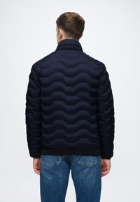 Ναυτική ζακέτα puffer με κυματιστό ραφτό σχέδιο, ψηλό γιακά και εφαρμόζοντες μανίκους, συνδυασμένη με ανοιχτό γαλάζιο τζιν.