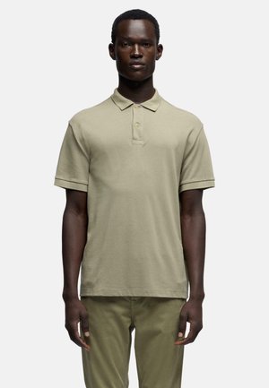 Mann trägt ein beiges Poloshirt mit kurzen Ärmeln und olivgrüne Hose, steht vor einem schlichten weißen Hintergrund.