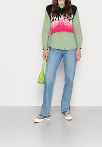 Person i en grön skjorta med en croppad tröja i svart och rosa flammmönster, ljusblå jeans och håller en liten grön väska.