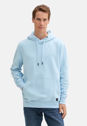 Man wearing light blue hoodie with front pocket and dark blue jeans, standing with one hand in pocket against plain white background.