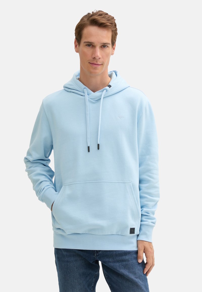 Uomo che indossa un felpa leggera blu con tasca frontale e jeans blu scuro, in piedi con una mano in tasca contro uno sfondo bianco semplice.