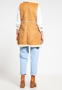 Brun faux-suede väst med fårskinnsdetaljer, lång design, baksidans knytning, bärs över en vit skjorta och ljusblå jeans. Bruna klackstövlar.