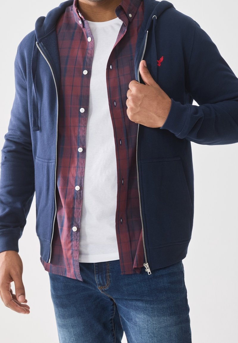 Homme portant un sweat à capuche bleu marine dézippé par-dessus une chemise à carreaux rouge boutonnée et un t-shirt blanc, associé à un jean bleu, ajustant la fermeture éclair du sweat à capuche.